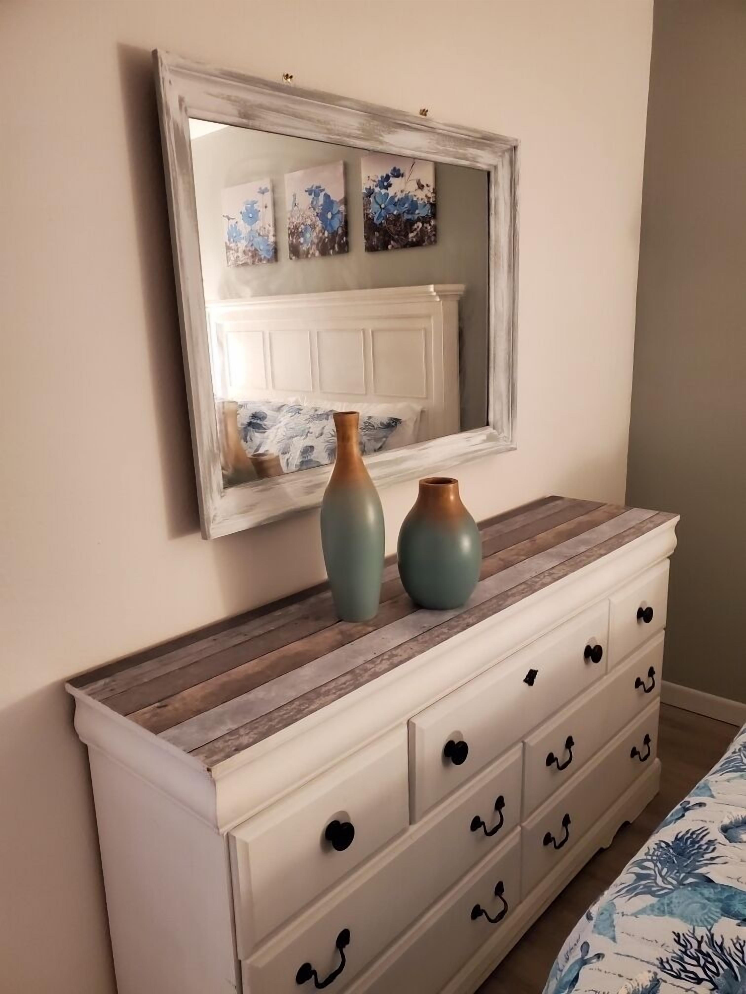 Ein Schlafzimmer mit einer weißen Kommode, zwei Vasen und einem Spiegel, der ein Bett mit Blumendekor reflektiert.