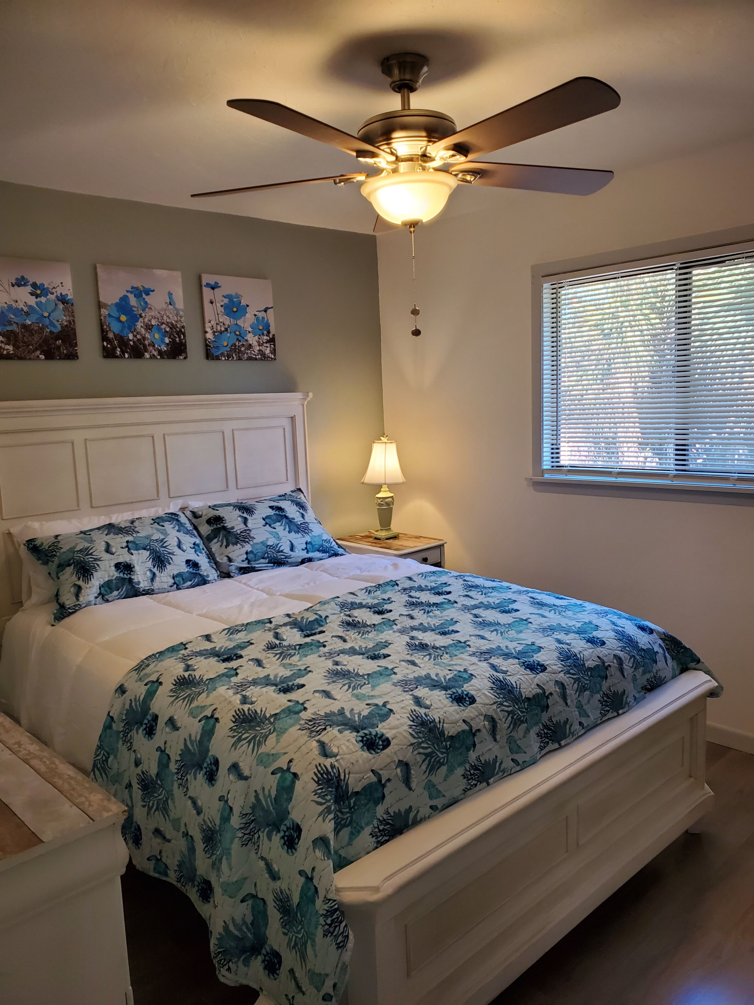 Une chambre à coucher avec un lit, un ventilateur de plafond, une fenêtre avec des stores et des œuvres d’art murales.