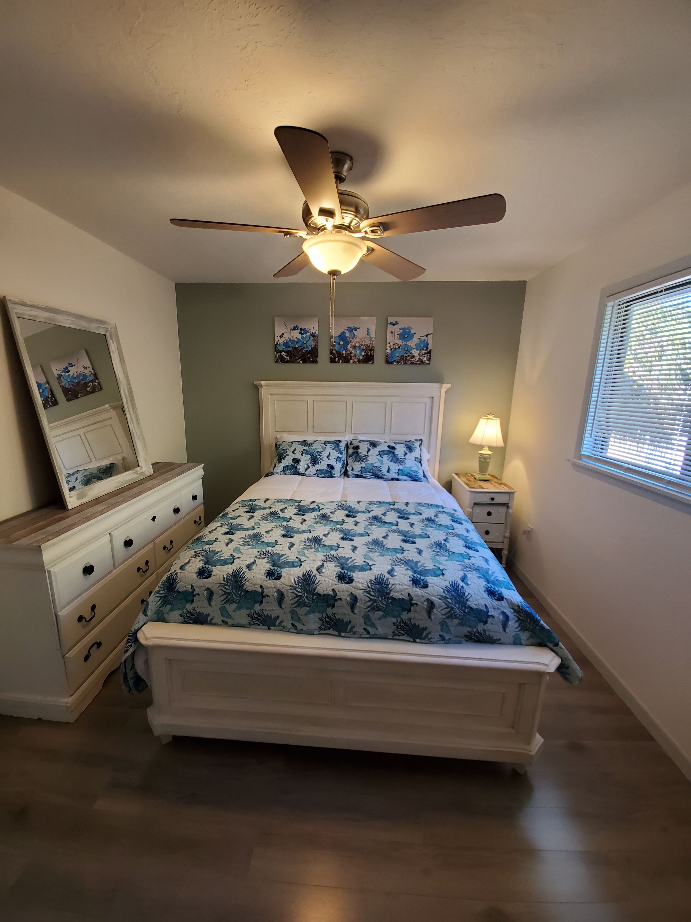 Ein Schlafzimmer mit Bett, Nachttisch, Lampe, Deckenventilator und Wandbild.