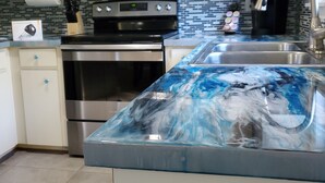 Une cuisine avec des meubles blancs, un four à micro-ondes en acier inoxydable, une cuisinière et un évier doté d’un plan de travail en marbre bleu.