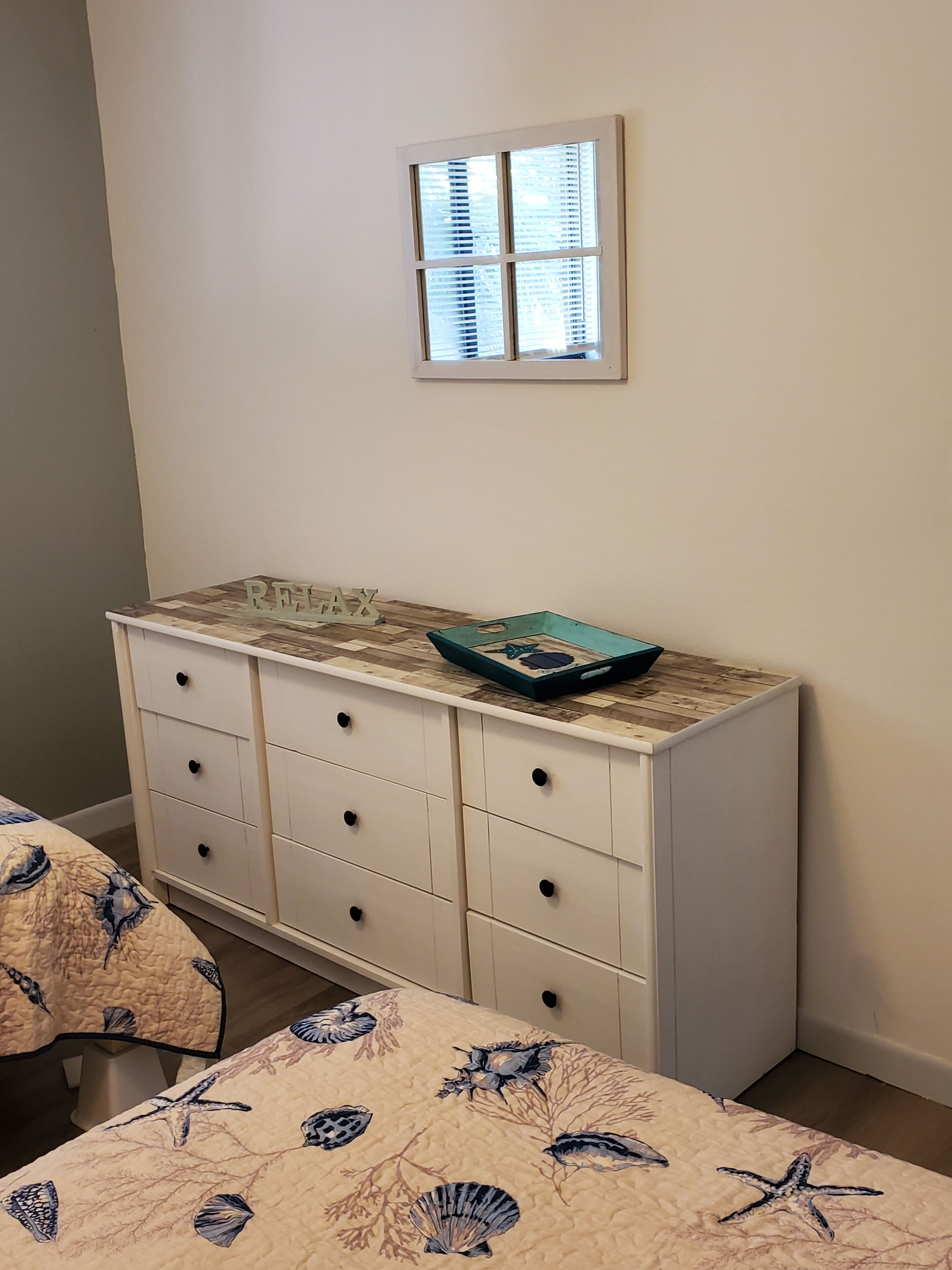 Un dormitorio con cama, cómoda, ventana y una mesita con un adorno.