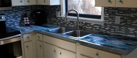 A kitchen with white cabinets, a black countertop, a stainless steel sink, and a window with a view of trees.