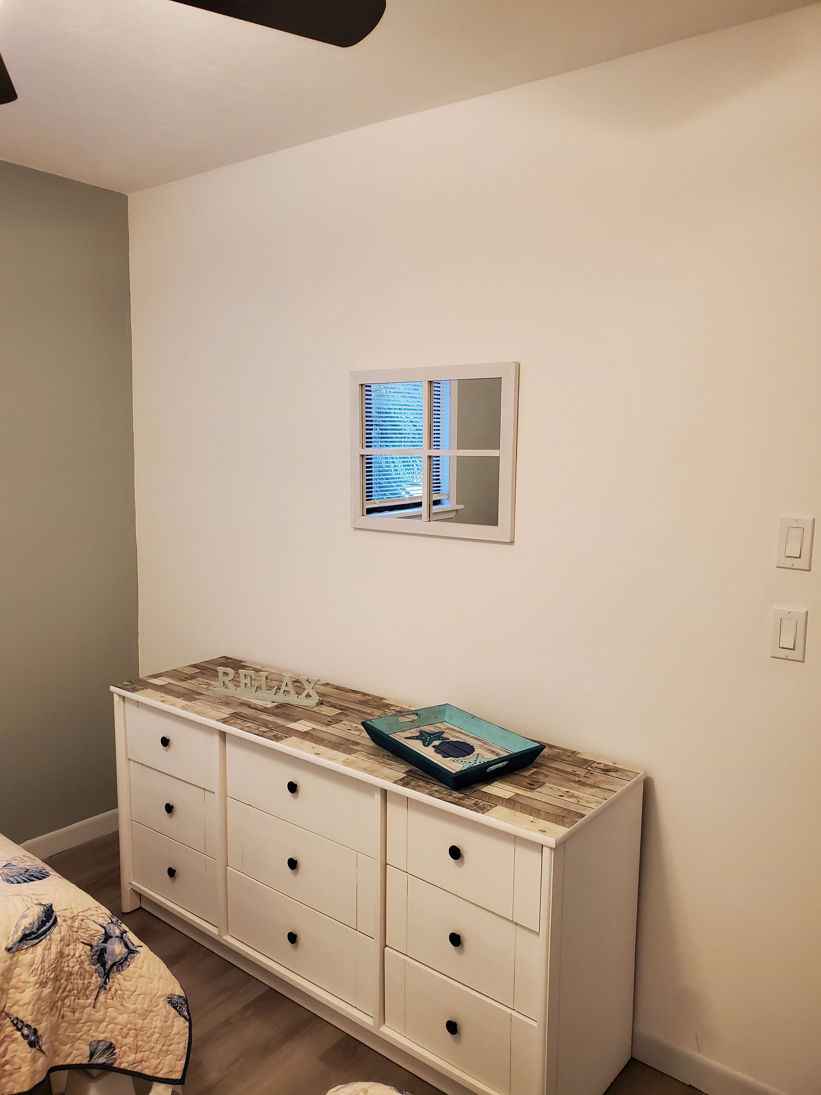A bedroom with a white dresser, a small window with blinds, and a bed with a quilt.