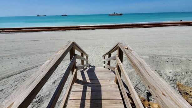 Une promenade en bois qui mène à une plage bordée d’une eau turquoise et transparente, avec des navires visibles au loin.