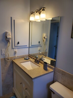 Een badkamer met een wastafel, een opbergmeubel en een toilet. Er zijn haardrogers aan de muur bevestigd, een spiegel boven de wastafel en een handdoekhouder.