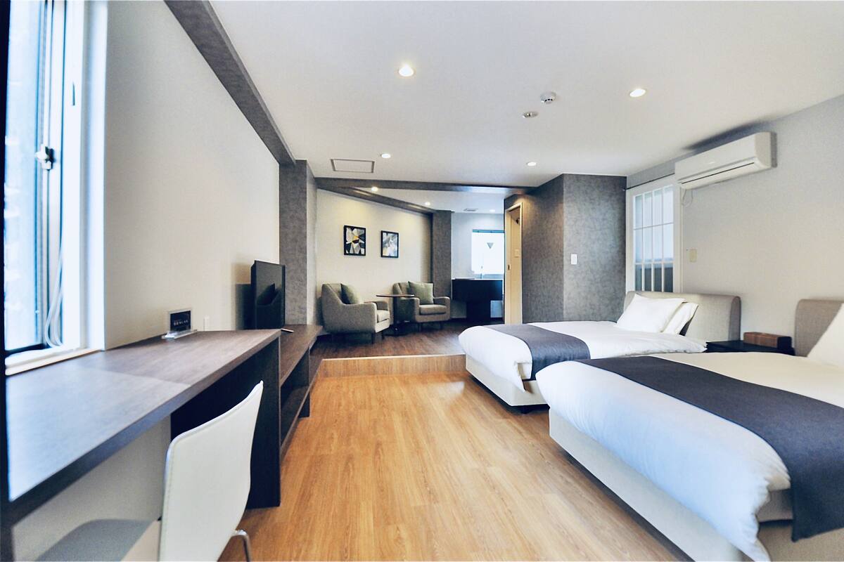 A modern hotel room with a desk, two beds, a seating area, and a large window.