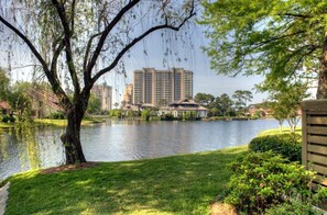 A serene lakeside view with a large tree in the foreground, a gazebo, and high-rise buildings in the background.
