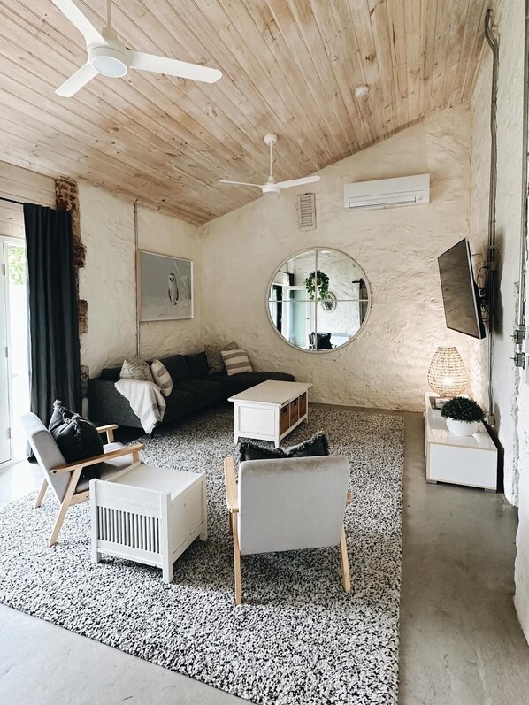A cozy living room with a wooden ceiling, a round mirror, a white sofa, a white coffee table, and a TV mounted on the wall.