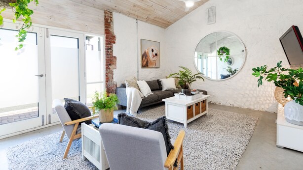 A modern living room with a grey sofa, white coffee table, and a large mirror. There are potted plants and a ceiling fan. - The Kings Stables at Burnside (Burnside)