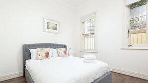 A neatly made bed with white linens and decorative pillows, a grey upholstered headboard, and a window with a view of a stone wall and greenery. - Stunning Sydney Home 7 (Millers Point)