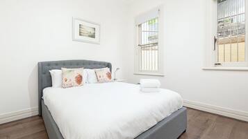 A neatly made bed with white linens and decorative pillows, a grey upholstered headboard, and a window with a view of a stone wall and greenery.
