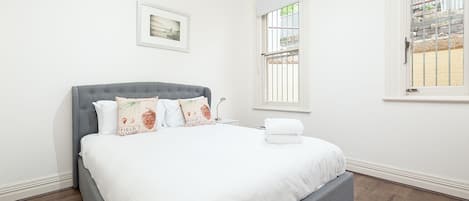 A neatly made bed with white linens and decorative pillows, a grey upholstered headboard, and a window with a view of a stone wall and greenery.