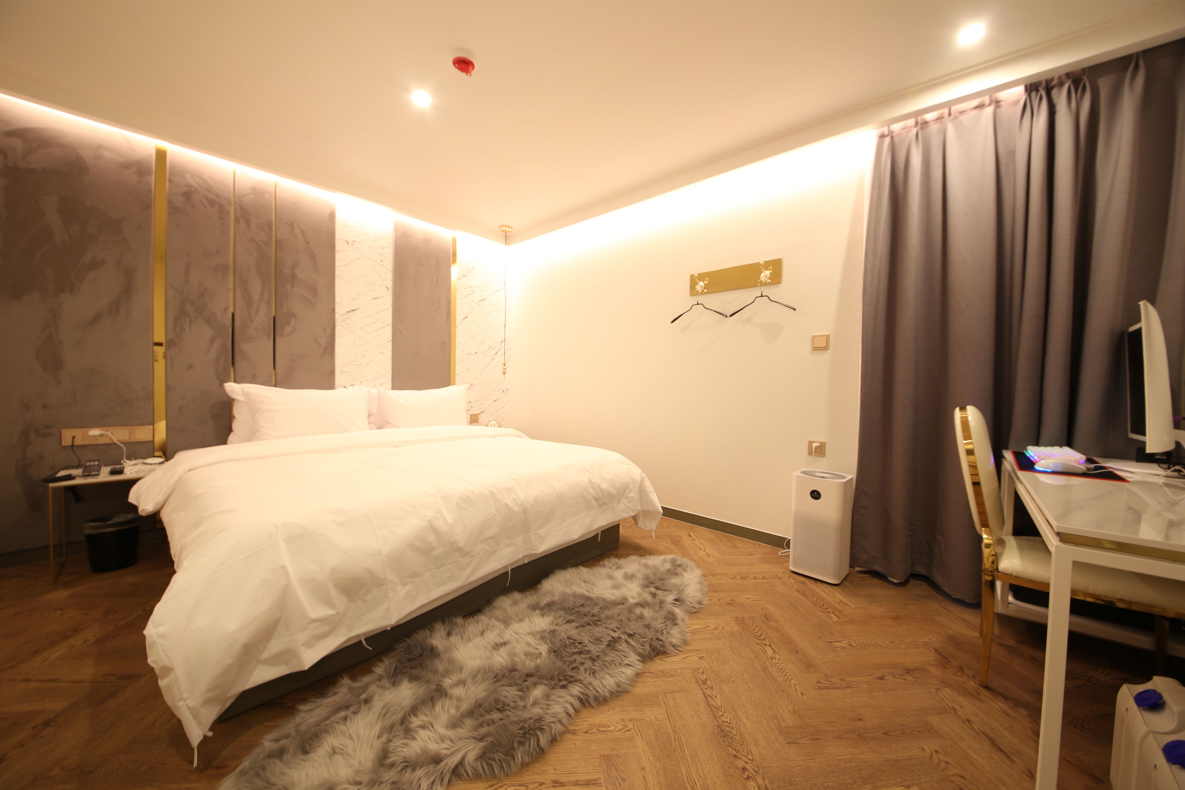 A modern bedroom with a large bed, a desk with a computer, and a small trash bin.