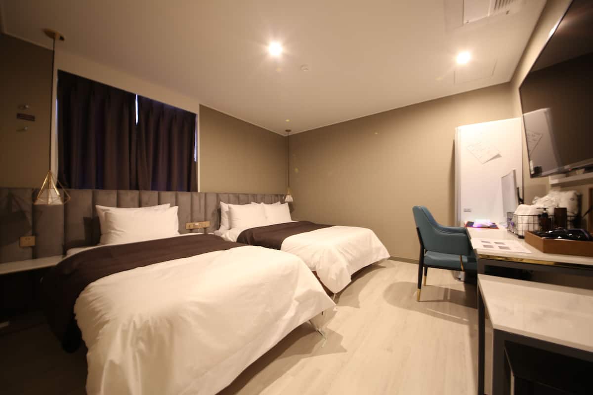 A hotel room with two beds, a desk with a chair, a large window with curtains, and a flat-screen TV mounted on the wall.