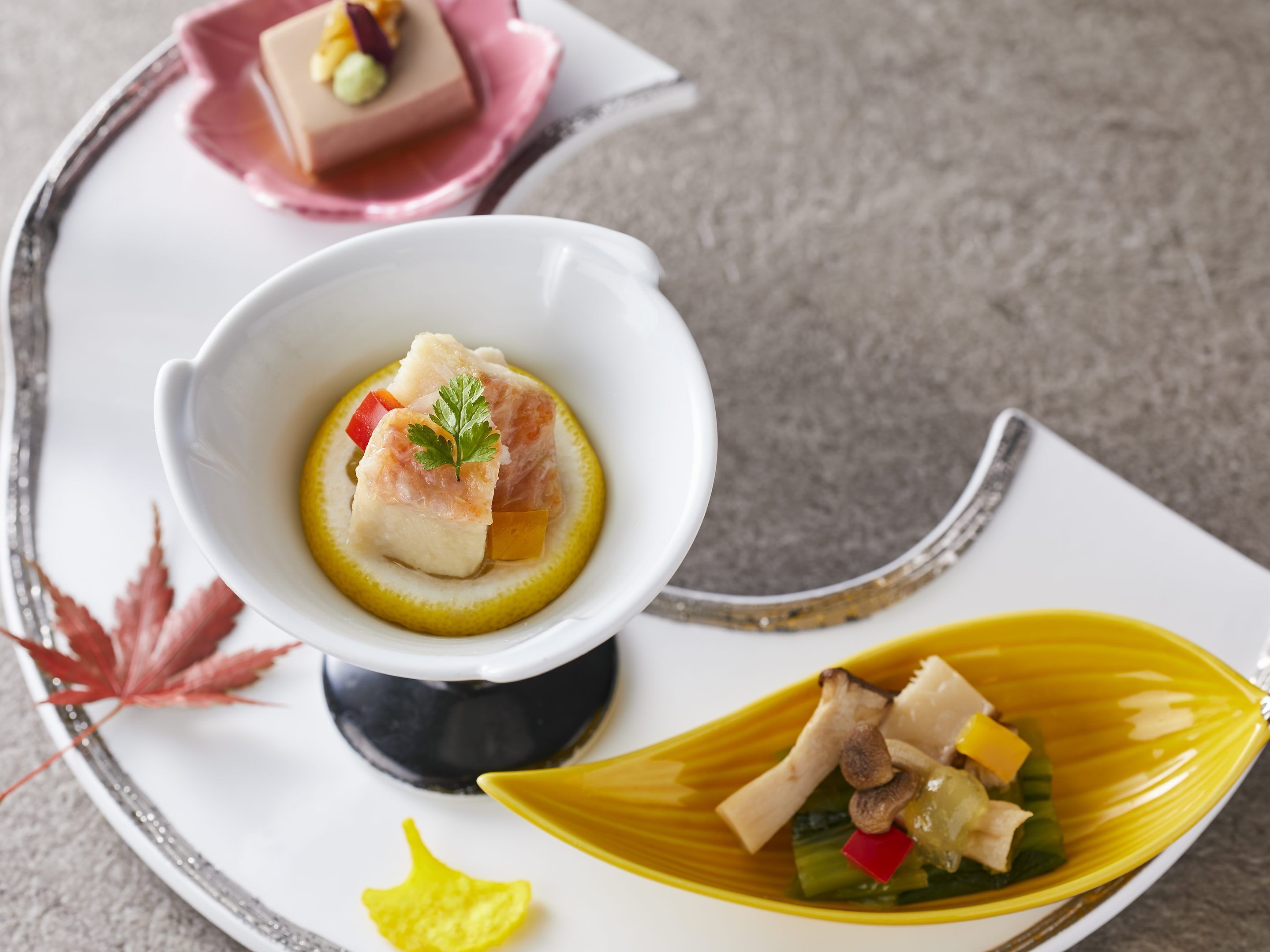 A plate with a white bowl containing a dish with fish and vegetables, a yellow leaf-shaped plate with more food, and a pink plate with a dessert.