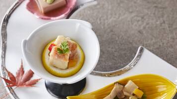 A plate with a white bowl containing a dish with fish and vegetables, a yellow leaf-shaped plate with more food, and a pink plate with a dessert.