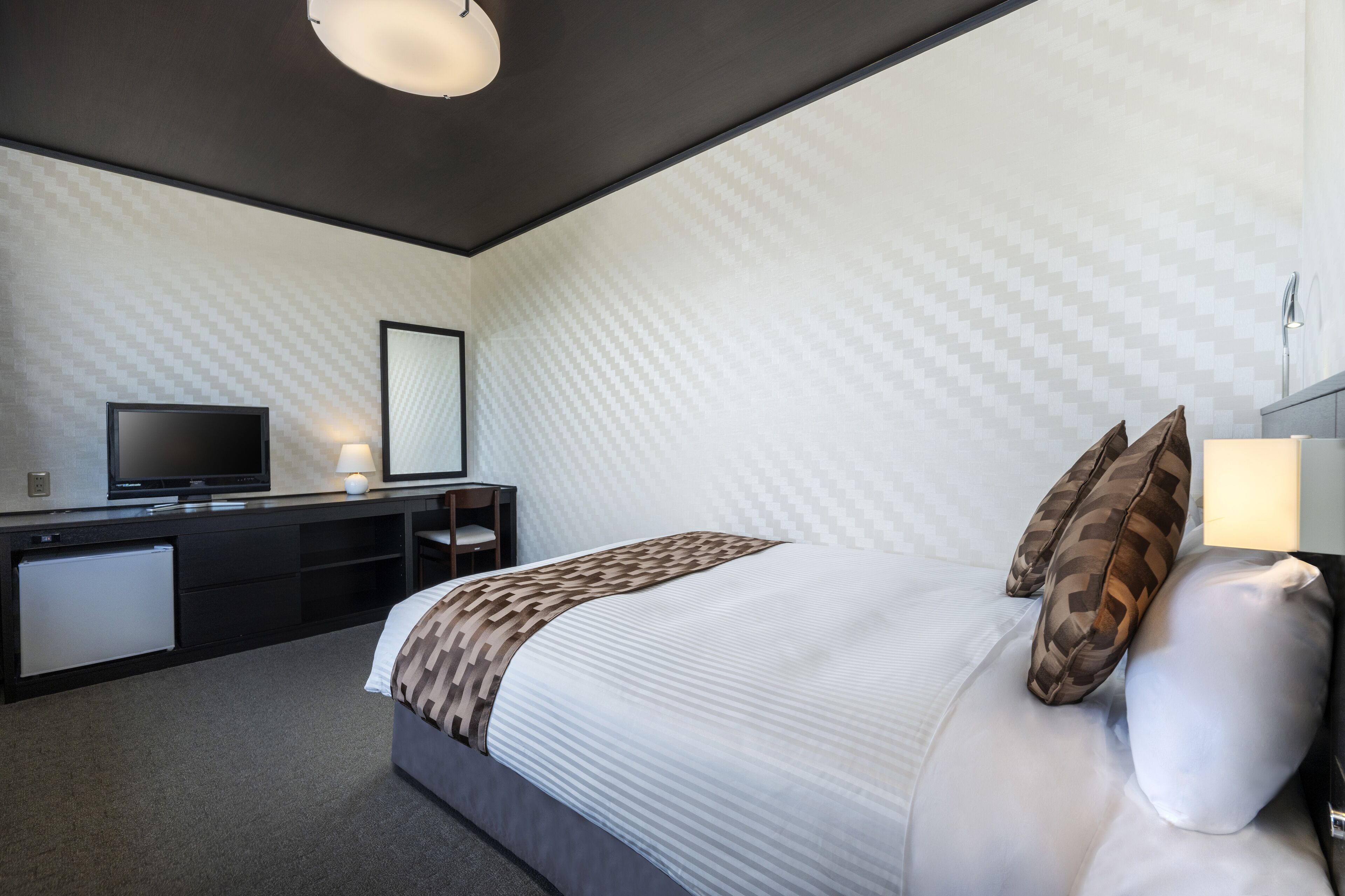 Quarto de hotel moderno com uma cama grande, uma televisão de tela plana, uma escrivaninha com abajur e um pequeno refrigerador.