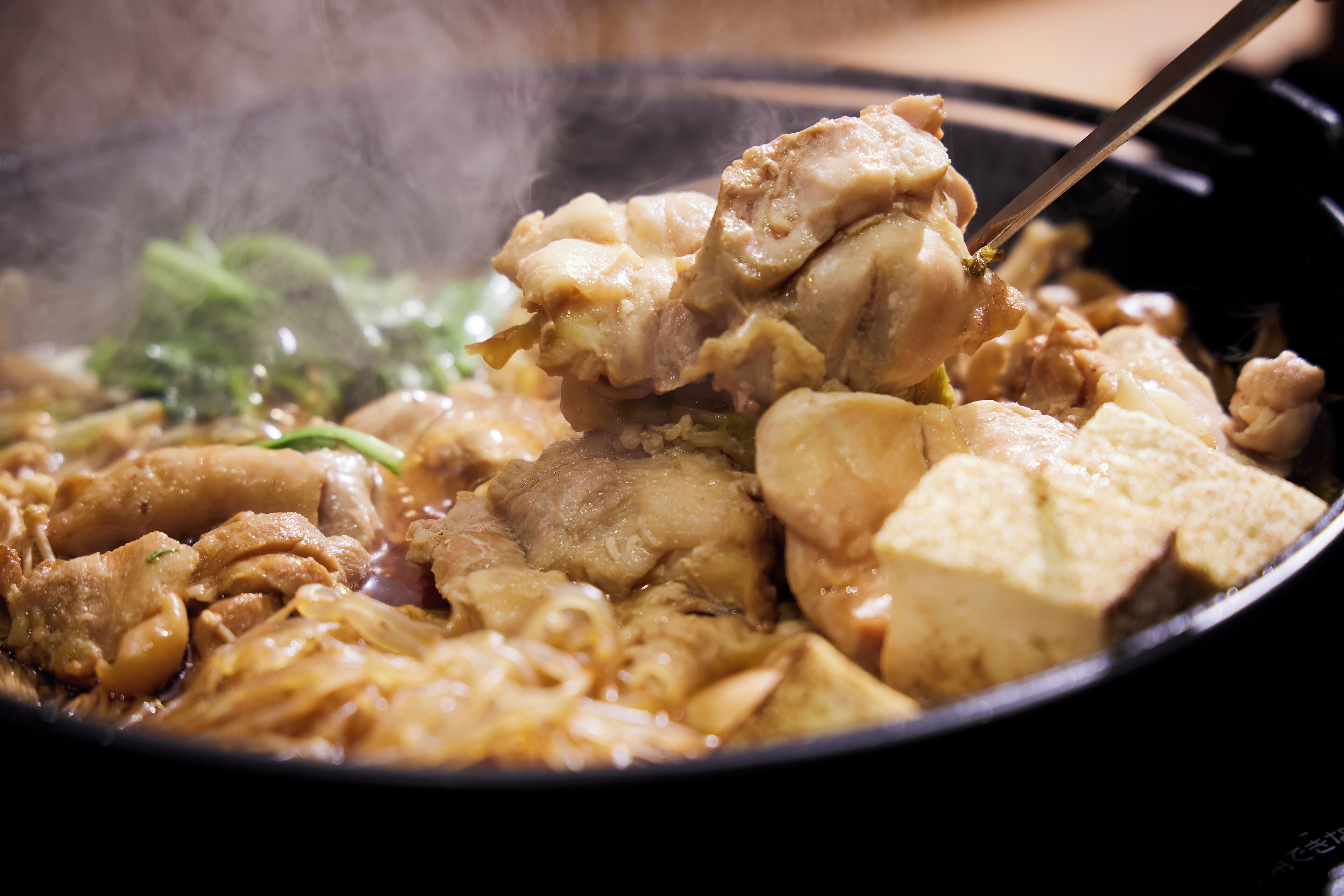 A close-up of a hot dish with various ingredients, including meat and vegetables, being served with chopsticks.