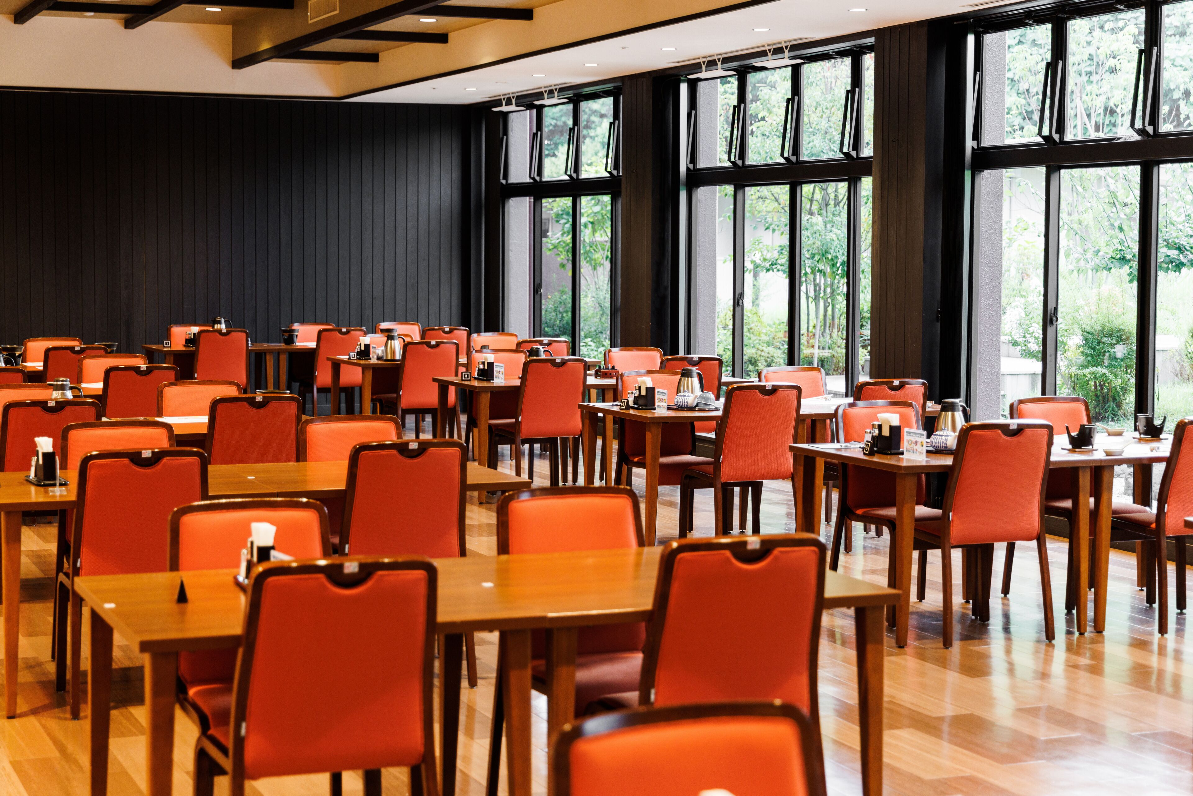 Un vaste espace dédié à la salle à manger, avec plusieurs tables et des chaises orange, de grandes fenêtres offrant une vue sur la verdure, et un mur sombre en arrière-plan.