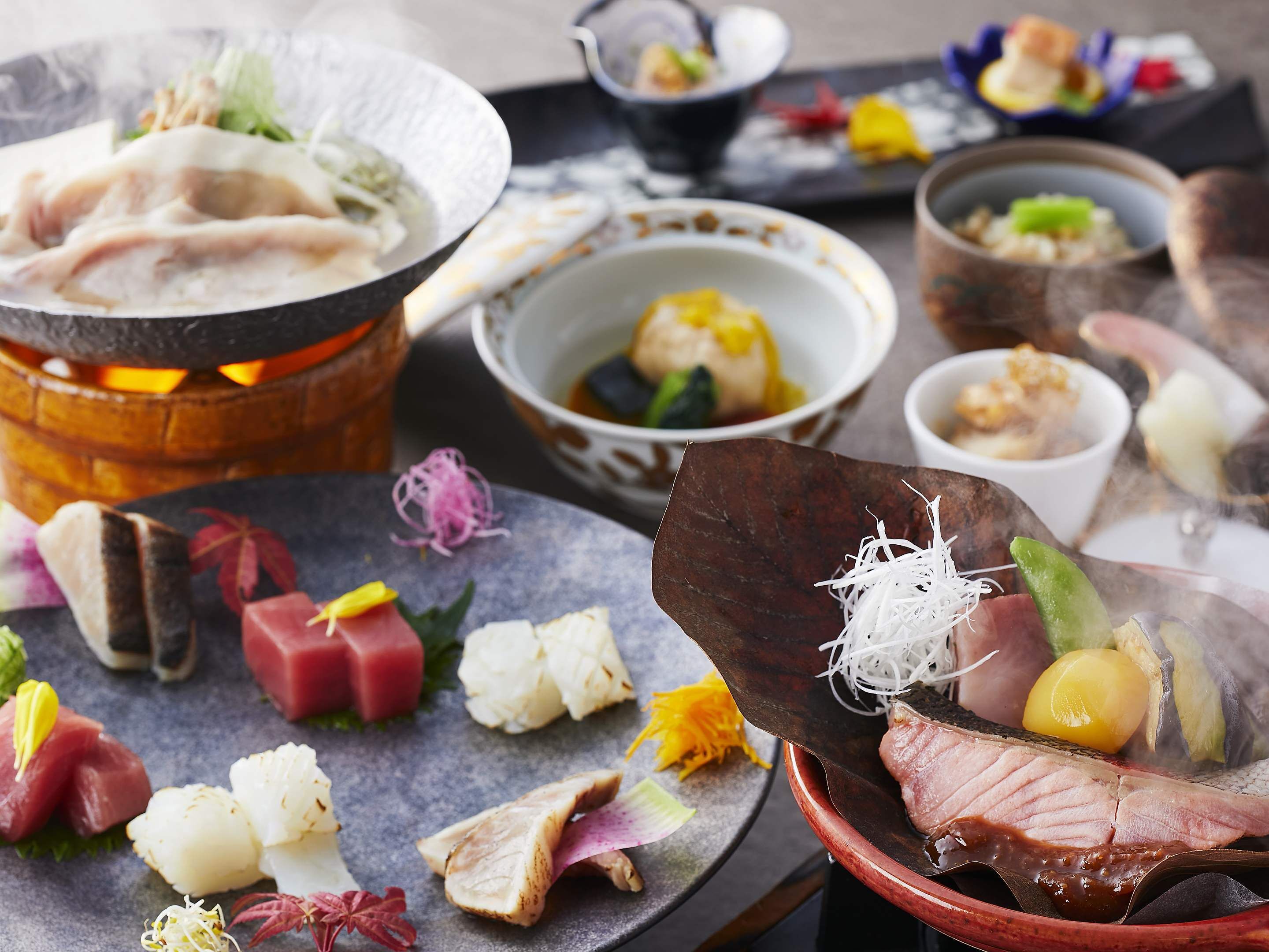 Une variété de plats de sushis et de sashimis sont disposés sur une table, avec un plat mijoté à portée de vue à l’arrière-plan.