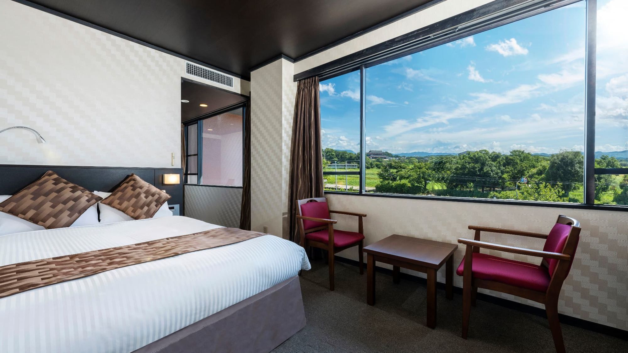 A hotel room with a bed, a table, two chairs, a window with a view of greenery, and a ceiling vent.