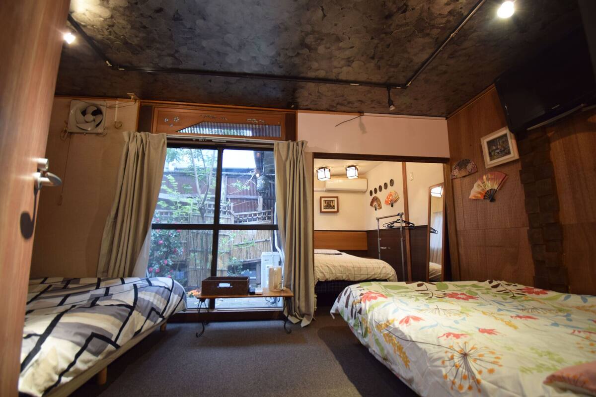 A room with two beds, a window with curtains, a wooden ceiling, and a small table with a chair.