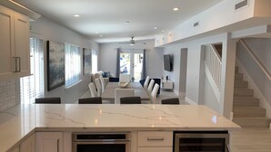 Cozinha moderna com ilha central, forno embutido e máquina de lavar louça. Possui área de jantar com mesa e cadeiras, e escada que leva a um andar superior.