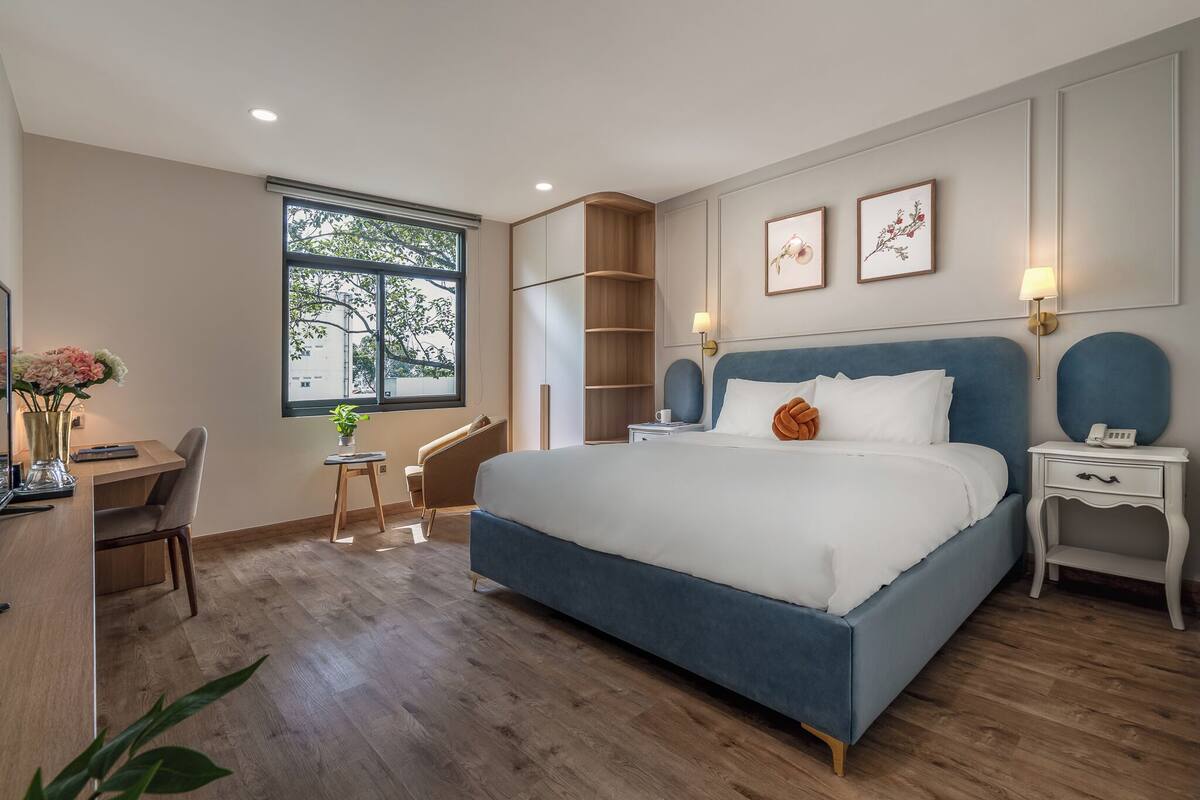 Une chambre avec un grand lit, des tables de chevet, un bureau et une chaise. Il y a une fenêtre donnant sur des arbres et une lampe sur la table de chevet.