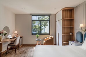 Ein modernes Schlafzimmer mit Schreibtisch aus Holz, einem Laptop, einem Stuhl, einem Nachttisch mit Lampe und einem großen Fenster mit Blick auf Bäume und Gebäude.
