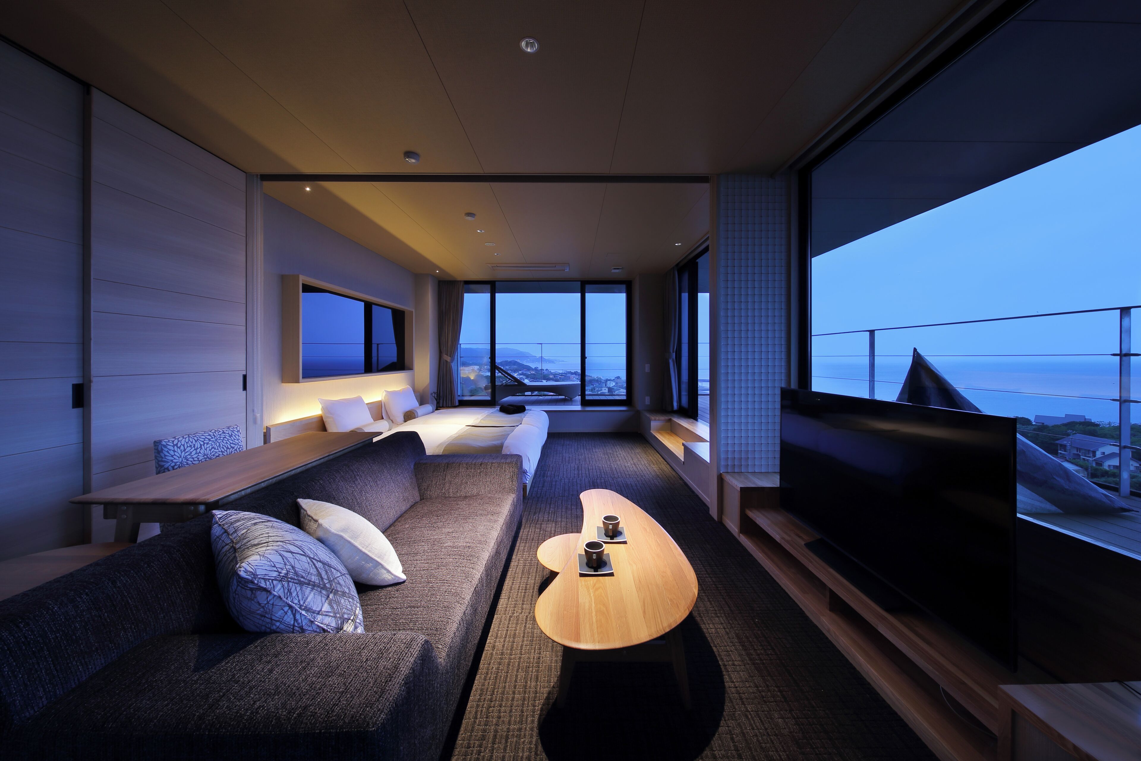 A modern hotel room with a large TV, a sofa, a coffee table, and a view of the city.