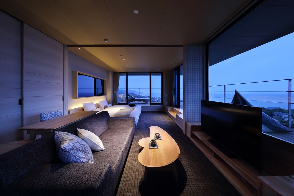 A modern hotel room with a large TV, a sofa, a coffee table, and a view of the cityscape.