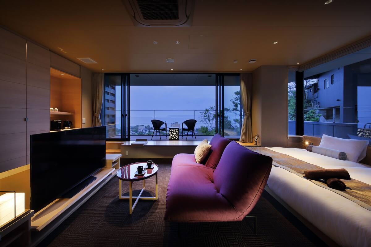A modern hotel room with a large flat-screen TV, a sofa, a bed, and a balcony with chairs.