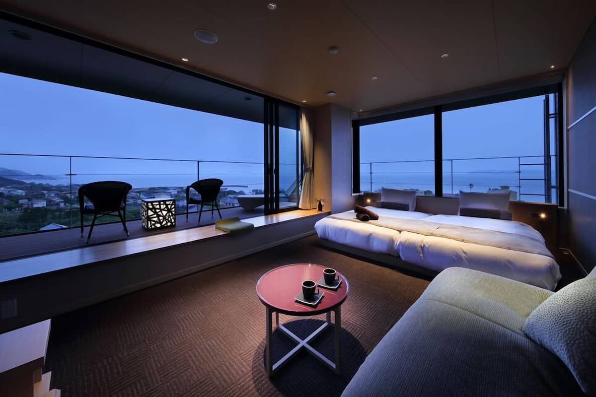 A modern hotel room with a large balcony, a sofa bed, a small round table, and a view of the cityscape.