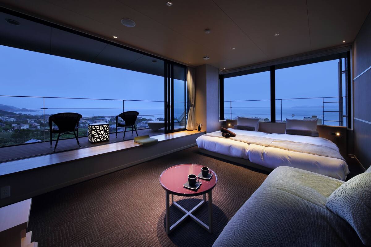 A modern hotel room with a large bed, a sofa, a small table with two cups, and a view of the cityscape through large windows.