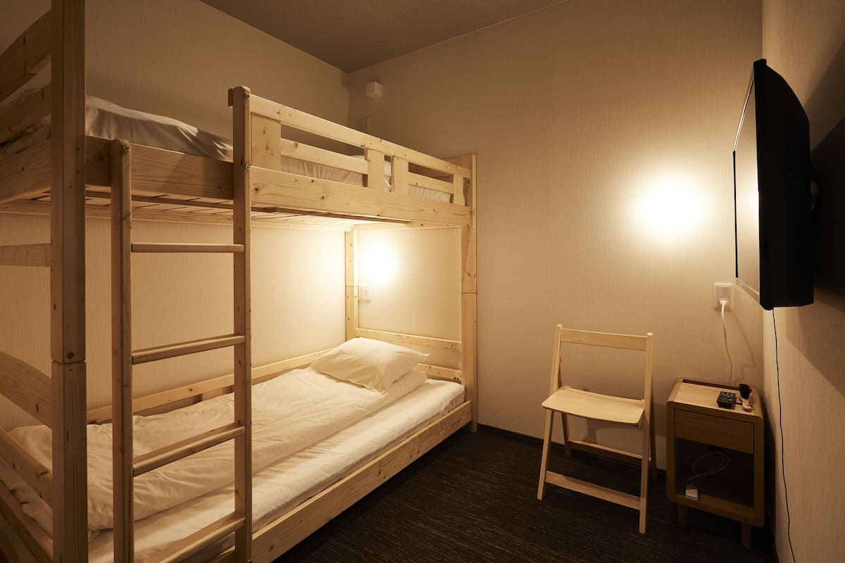 Habitación con literas, un escritorio, una silla, un televisor montado en la pared y un enchufe.