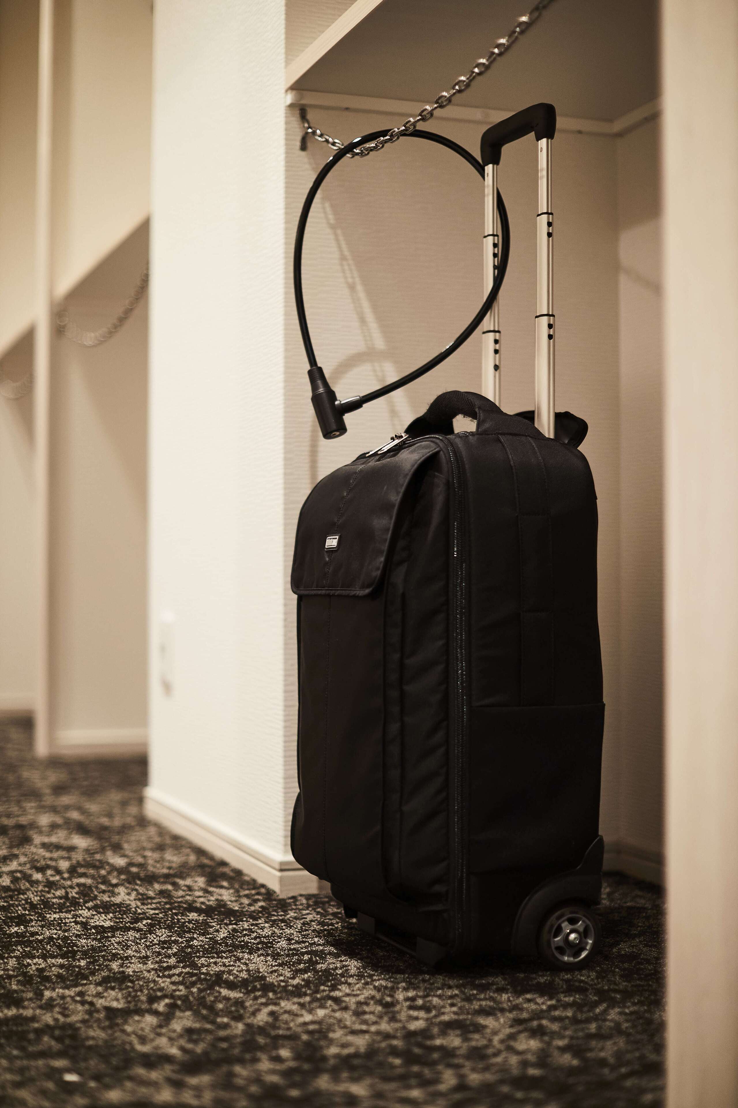 A black rolling suitcase with wheels and a handle, leaning against a wall in a hallway with carpeted flooring.