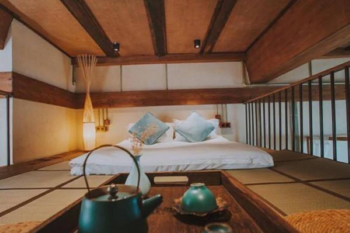 A traditional Japanese-style bedroom with tatami flooring, a low wooden ceiling, and a bed with pillows and a blanket. There is a wooden tray with a green teapot and two cups on the floor.