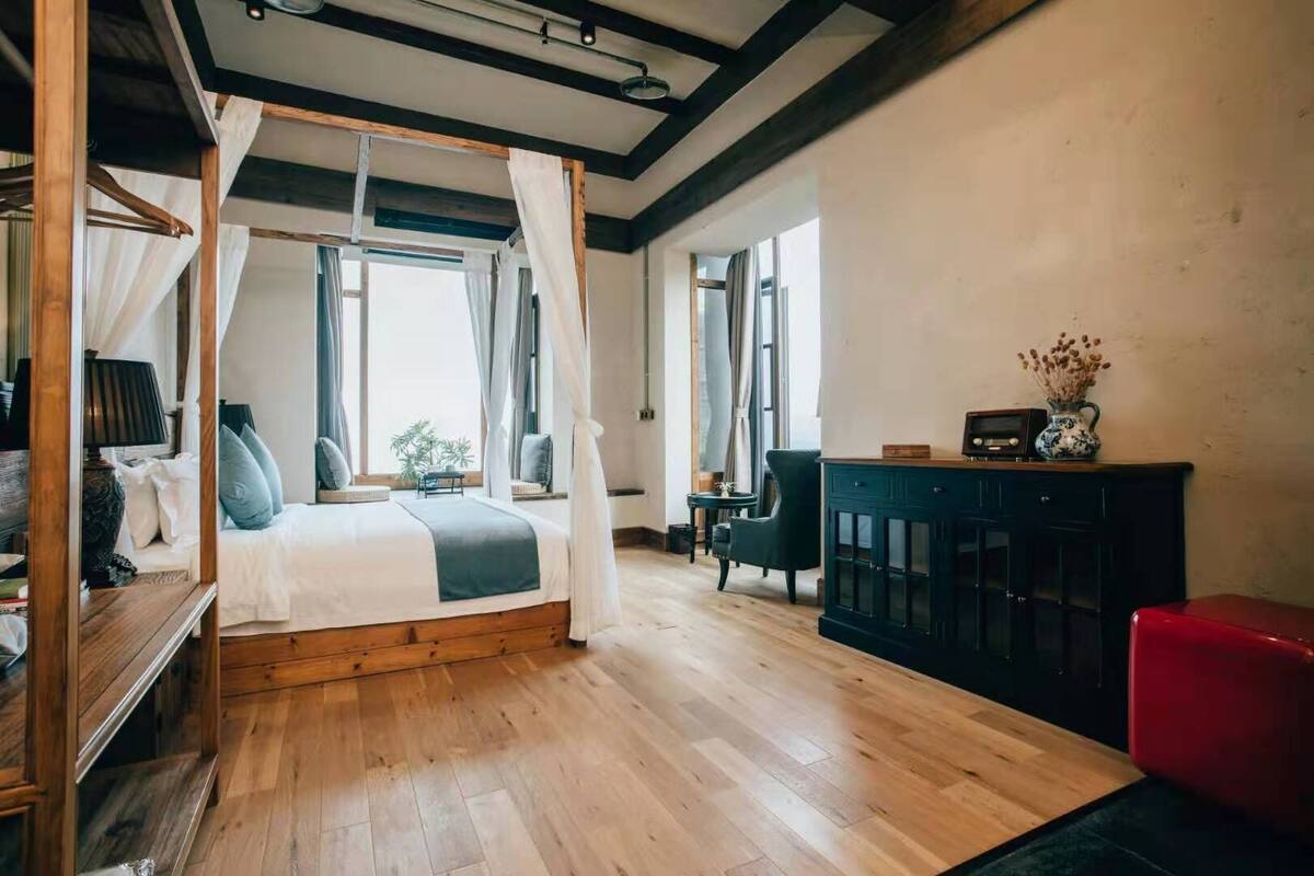 A bedroom with a four-poster bed, a wooden floor, a dark-colored cabinet, and a red chair.