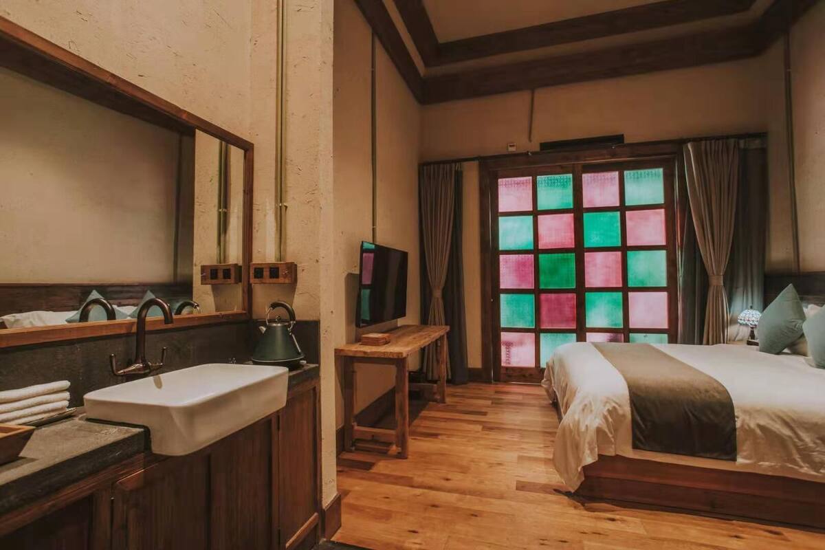 A room with a wooden floor, a bed with a white bedspread, a wooden nightstand with a television, a wooden dresser with a sink, and a window with colorful panes.