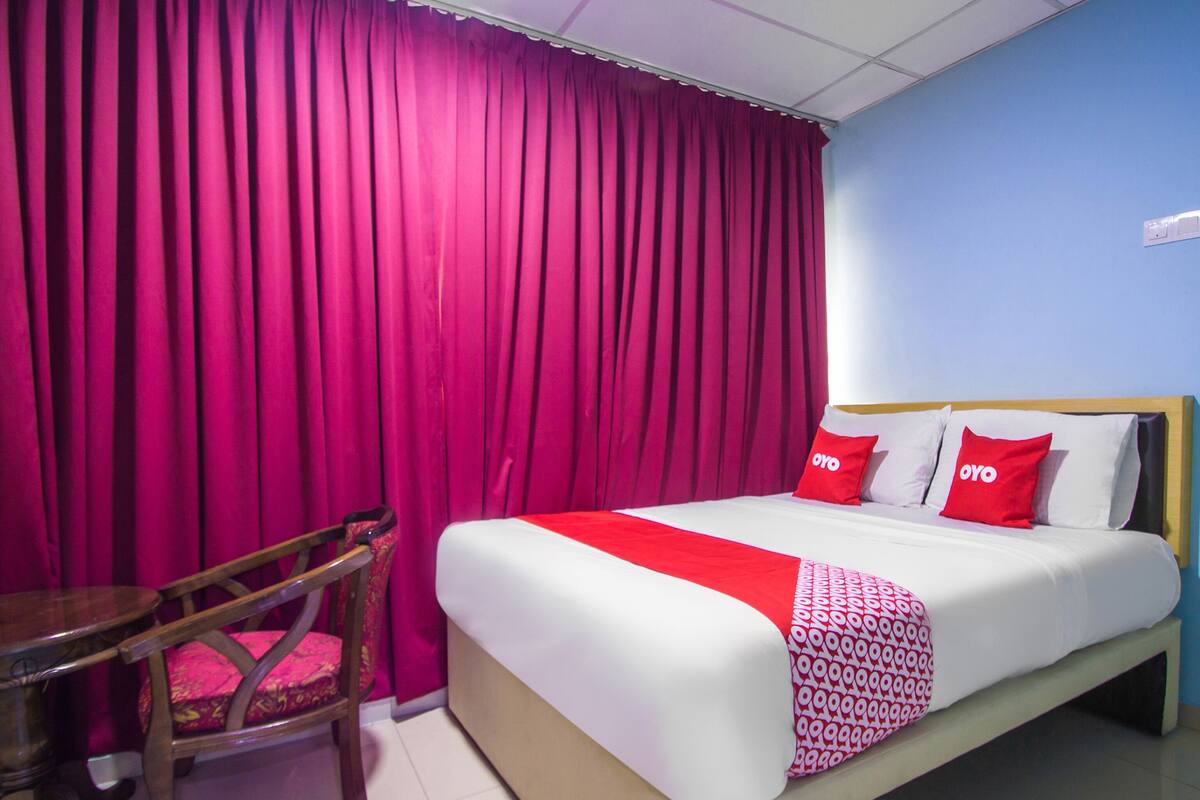 A hotel room with a bed, a wooden chair, and a large window with curtains.