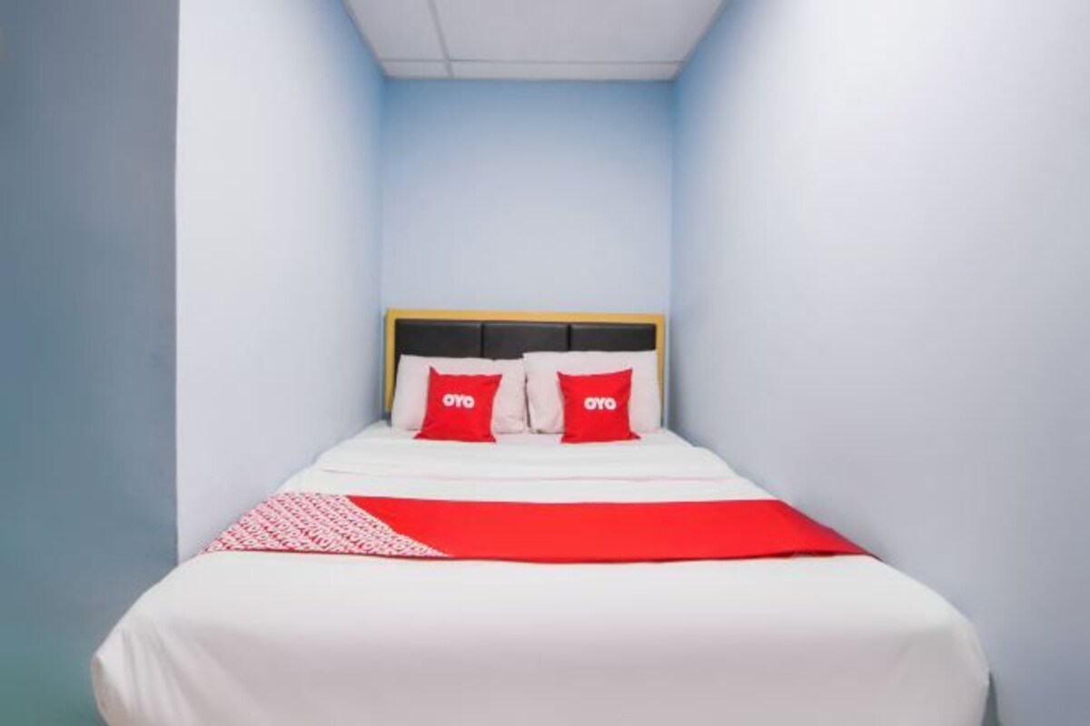 A narrow hotel room with a single bed, a wooden headboard, and two red pillows.