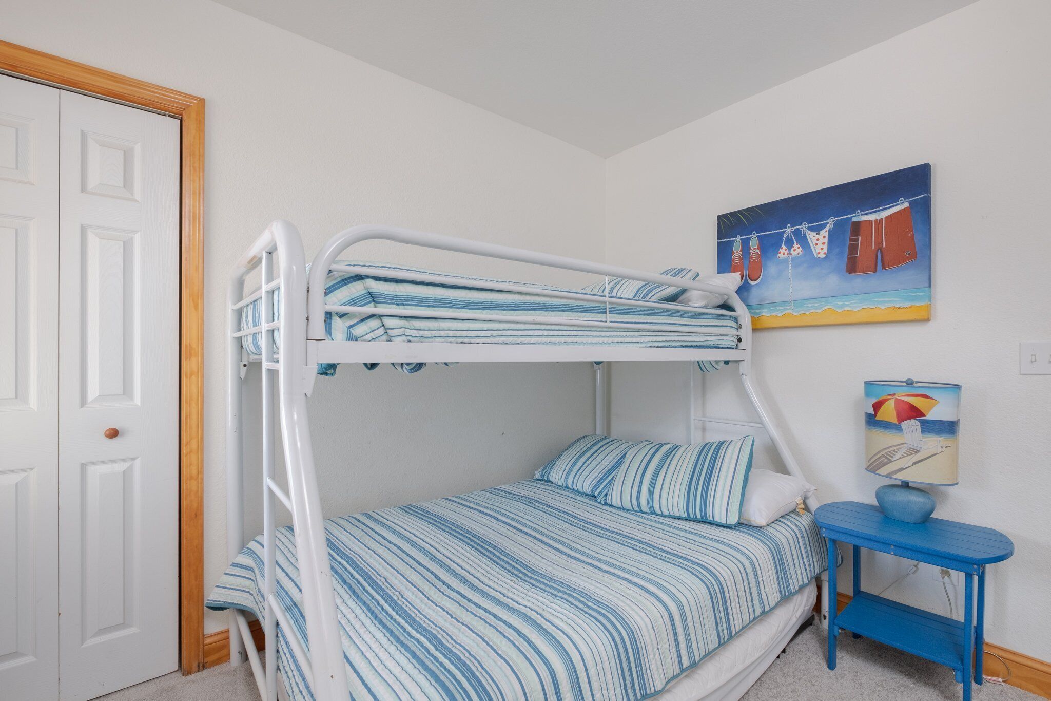 Ein Etagenbett mit gestreifter Bettwäsche, ein kleiner Tisch mit Lampe sowie ein Bild mit Strandkleidung an der Wand.
