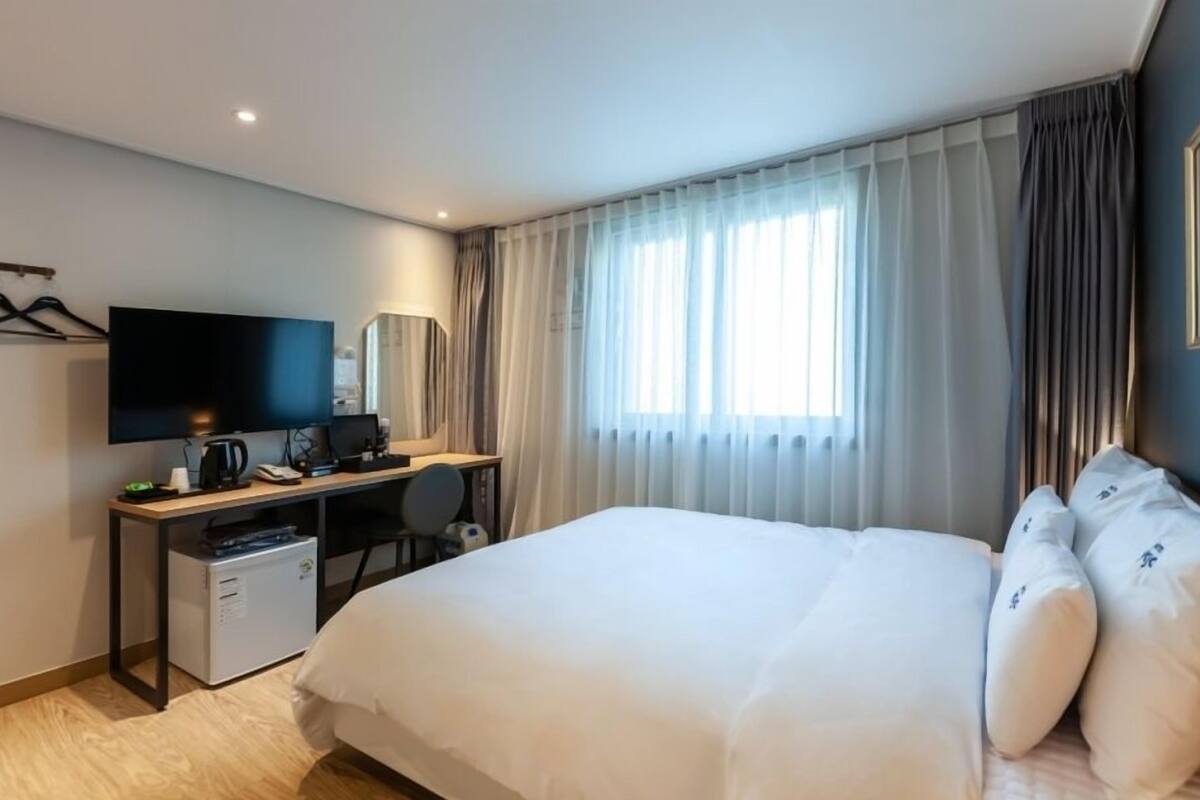 A hotel room with a bed, a desk with a computer, a TV, and a small kitchenette.