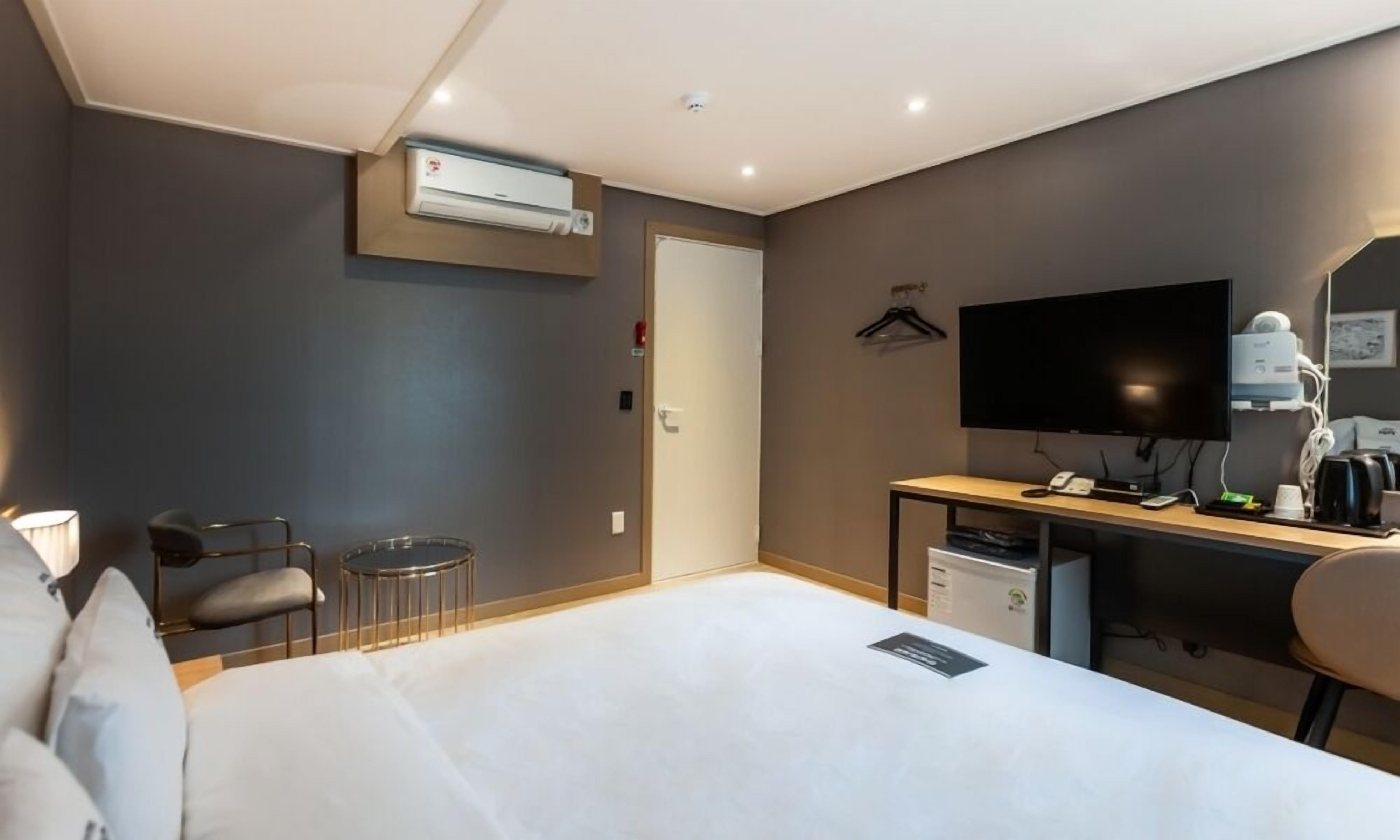 A hotel room with a bed, a desk with a computer, a chair, a TV, and an air conditioning unit.