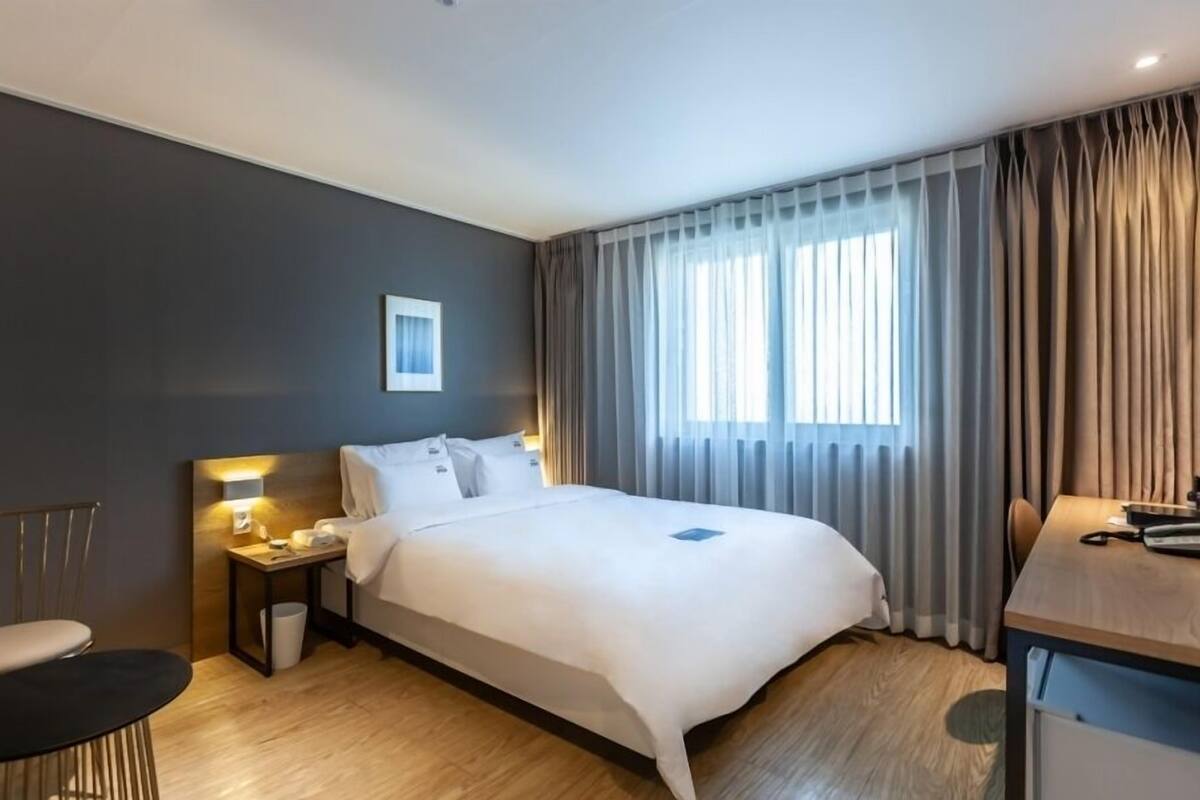 A hotel room with a bed, bedside table, lamp, chair, and a desk with a computer.