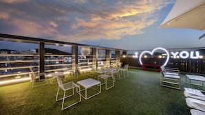 An outdoor seating area with white metal chairs and tables, a 'I ♥ YOU' sign, and a view of the cityscape at dusk. - YAJA Hotel Tongyeong Ferry Terminal Branch (Tongyeong)