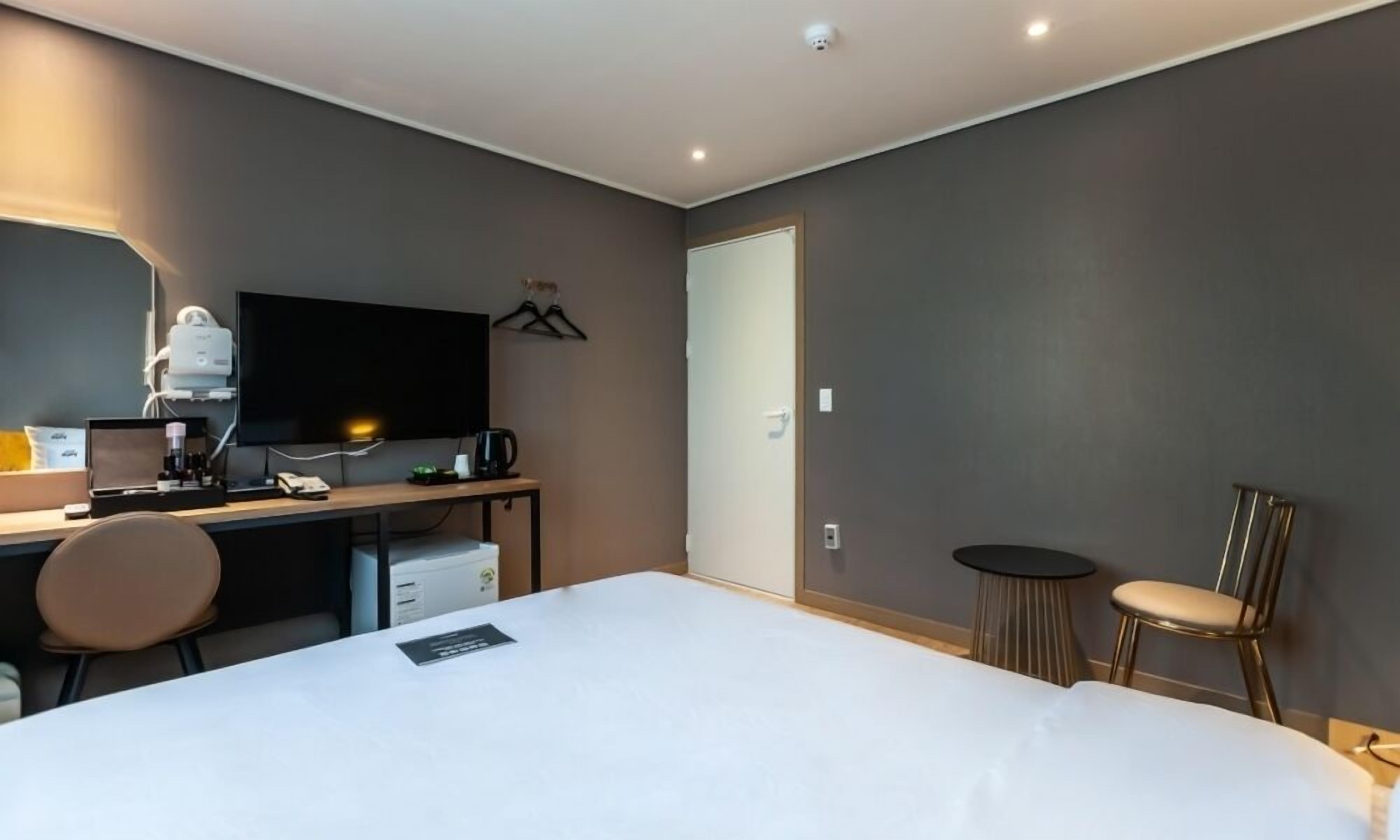 A modern hotel room with a large bed, a desk with a computer, a chair, a TV, and a small table with a lamp.