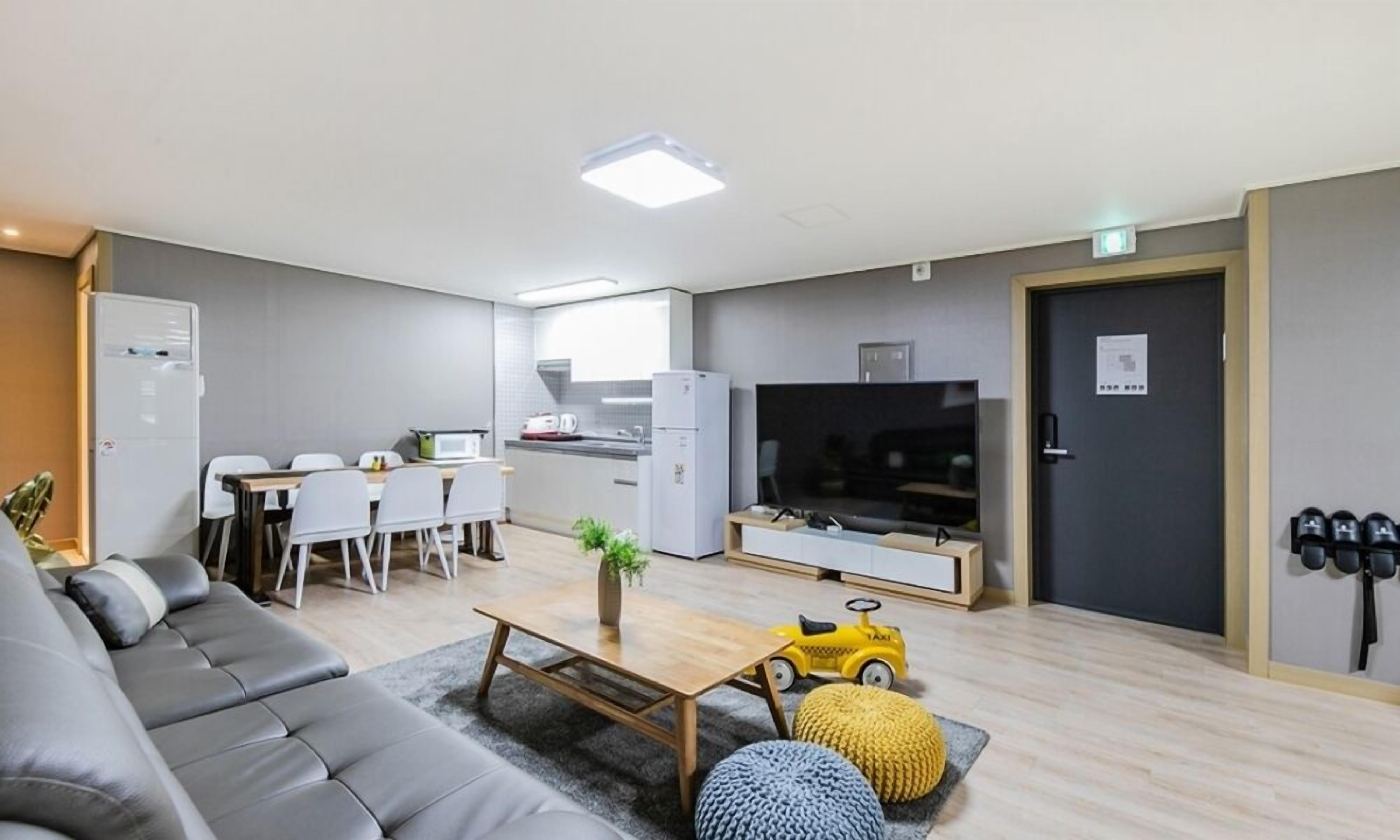 A modern living room with a grey sofa, a wooden coffee table, and a TV. There is a kitchen area with a fridge and a microwave.