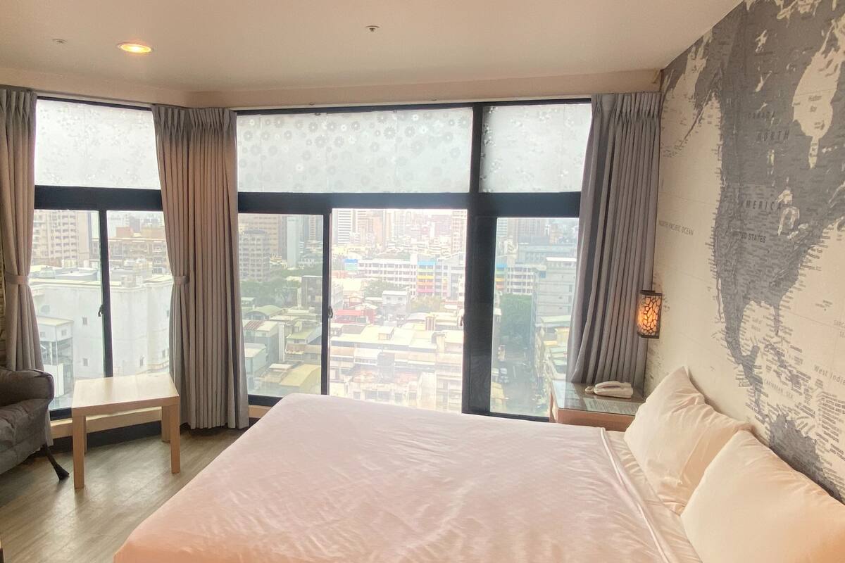 A hotel room with a large bed, a desk, and a view of a cityscape through large windows.