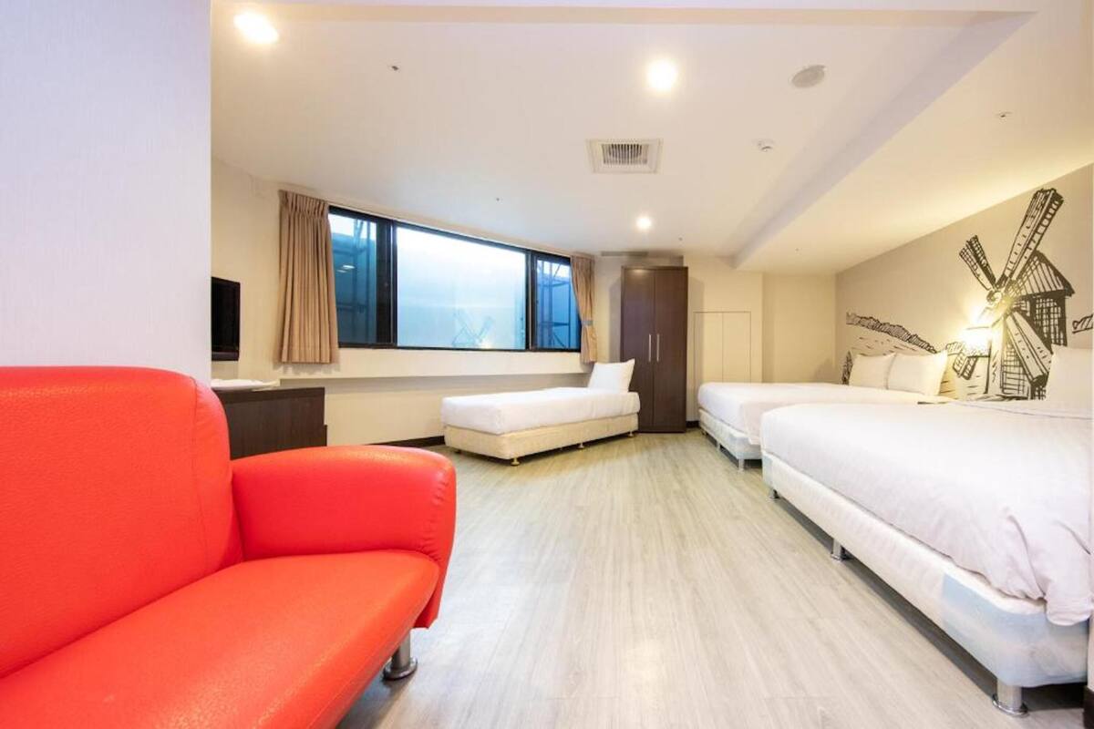A modern hotel room with a large window, a red sofa, two beds, and a windmill wall art.